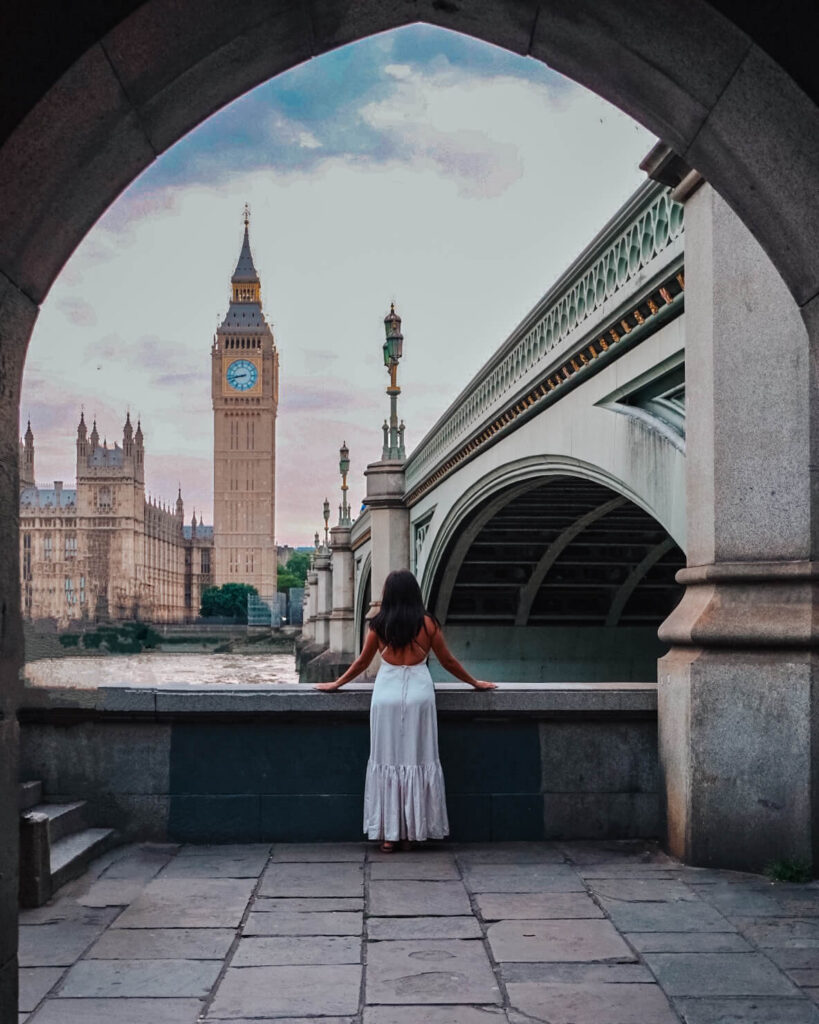 A woman in a white dress stands at an archway, overlooking the River Thames with Big Ben and Westminster Bridge in the background. The sky has hues of pink and purple, suggesting sunset.