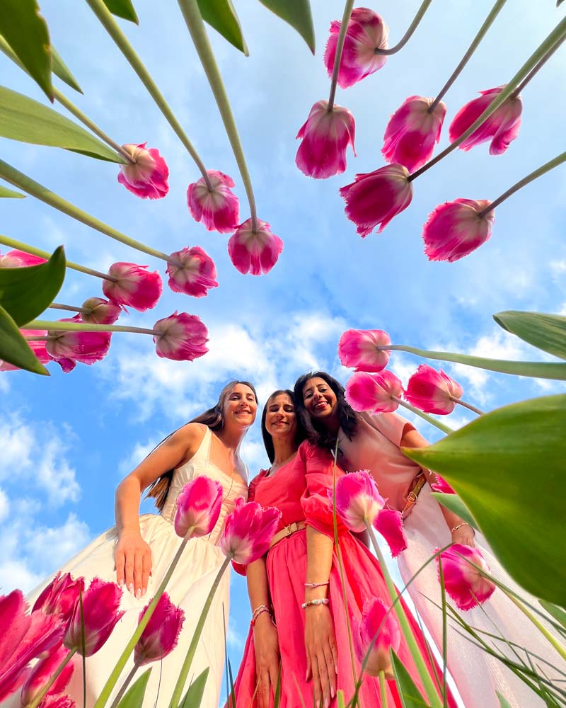 Three women smiling in a tulip field, captured from a low angle looking up through bright pink tulips toward a blue sky with scattered clouds.